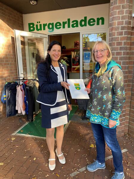 Samtgemeindebürgermeisterin Claudia Sommer überreicht Frau Ludwigs vor dem  Bürgerladen Nienhagen eine Ehrenurkunde.