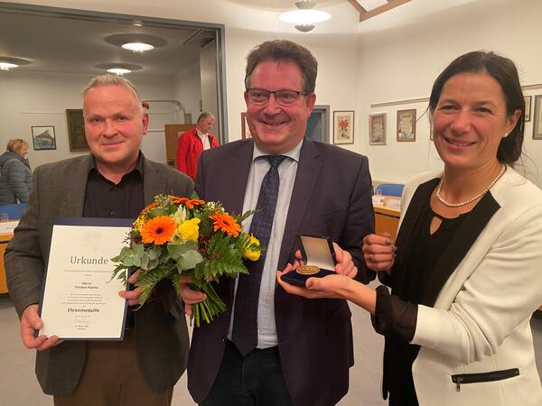 Bild vergrößern: Bürgermeister Harms steht in der Mitte mit einem Blumenstrauß, links von ihm steht Herr Hausknecht mit der Urkunde und rechts Frau Sommer mit der Medaille.