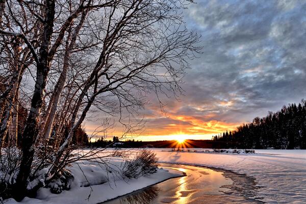 Eine verschneite Landschaft, durch die sich ein gefrorener Bach zieht, und im Hintergrund geht golden die Sonne unter.