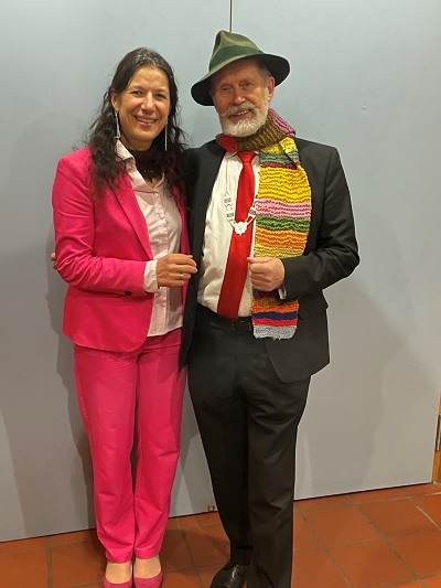 Frau Sommer im pinkfarbenen Anzug gratuliert Herrn Grube mit Amtskette, buntem Schal und Hut zur Ehrung.