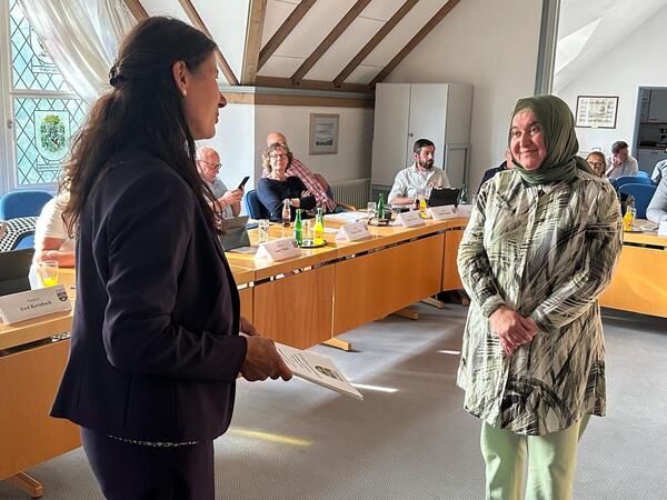 In einem hellen Konferenzraum &uuml;berreicht eine Frau im dunklen Hosenanzug einer l&auml;chelnden Frau mit Kopftuch und gemustertem Oberteil eine Urkunde. Im Hintergrund sitzen mehrere Personen an einem Konferenztisch mit Namensschildern, Getr&auml;nken und Laptops. Die Atmosph&auml;re wirkt feierlich und respektvoll.