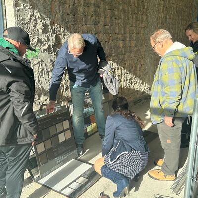 Bild vergr&ouml;&szlig;ern: Auf dem Boden stehen Muster von Fliesen, die von einer Gruppe Menschen begutachtet werden.