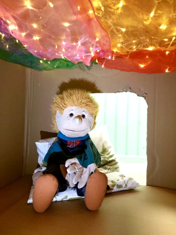 Bild vergr&ouml;&szlig;ern: Eine Stoffpupe sitzt in einem Karton unter einem bunten Dach aus Seident&uuml;chern.