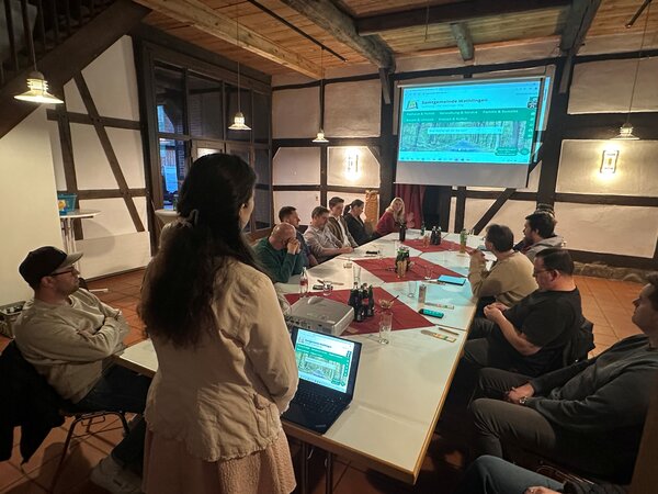 Bild vergr&ouml;&szlig;ern: Das Bild zeigt einen langen Tisch mit G�sten, die auf eine Pr�sentationsfl�che blicken.