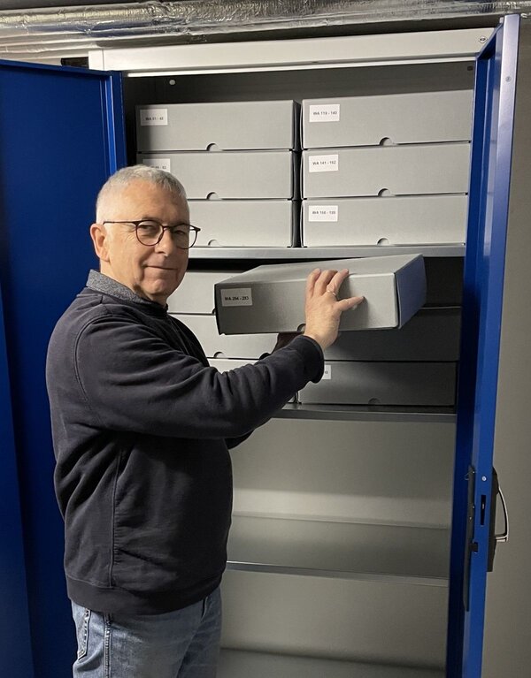 Herr Thunich steht vor einem Schrank, in dem viele graue K&auml;sten stehen und h&auml;lt eine Kiste in den H&auml;nden.
