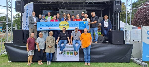 Bild vergr&ouml;&szlig;ern: Eine Gruppe von Menschen steht auf einer B�hne und h�lt ein Banner in den H�nden mit dem Schriftzug " Ein Projekt der Leader Region aller Fuhse Aue"